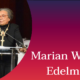 Marian Wright Edelman on stage with a drum set behind her. She is dressed in a black suit with a yellow collar and wearing a medal on her neck. There is a microphone stand in front of her and her mouth is above the microphone. Next to her is a curve of an orange, pink, and purple color palette with her name written in white.