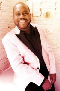 A black man with short hair and a trimmed beard smiles and stands at an angle wearing a whit tuxedo jacket with a black button down shirt.